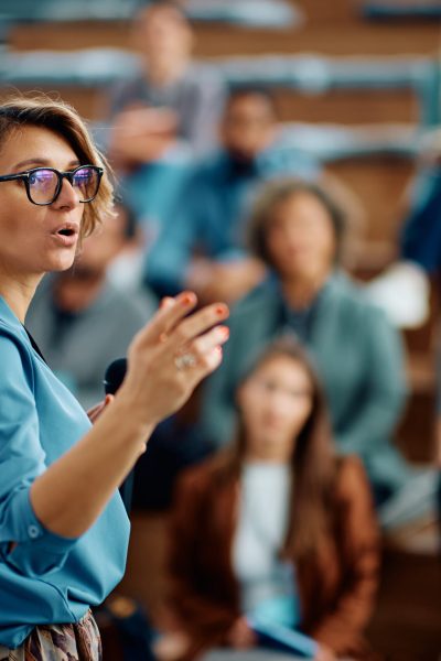 Mid adult businesswoman talking to group of seminar attendees in conference hall.